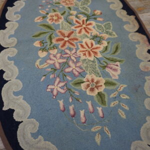 Oval light blue rug with a pink and green floral pattern and scrolling border on distressed wooden flooring.
