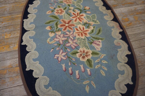 Oval light blue rug with a pink and green floral pattern and scrolling border on distressed wooden flooring.
