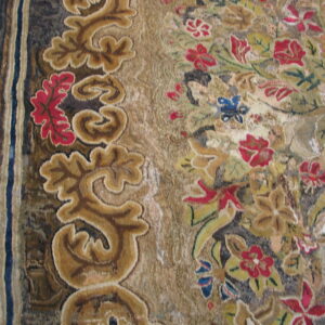 Close-up of a textured rug with a tan scroll border and multicolored floral pattern on hardwood floor.