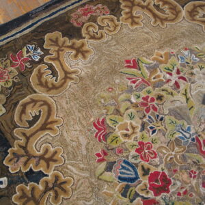 Textured, low-pile rug displaying intricate floral and baroque scroll patterns in muted tan, red, blue, and brown tones.