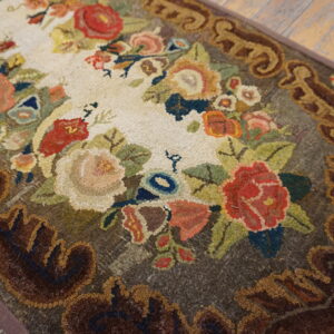 A looped pile rug with a colorful floral center and a dark brown scrolling border rests on wood planks.