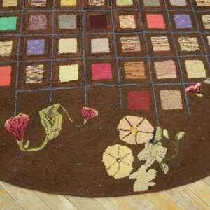 Textured brown rug with a colorful grid pattern and large floral motifs on the rounded edge set on wood flooring.