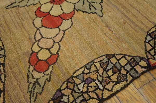 Low-pile hooked rug displays red, cream, and green floral motifs on a textured tan background over wood plank flooring.
