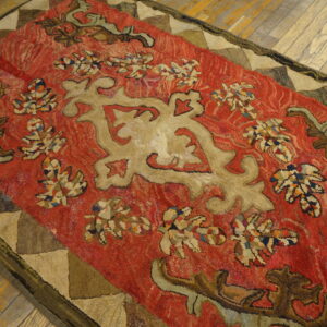 Rough texture red rug with tan scrollwork and a triangular patched border on worn wooden floorboards.