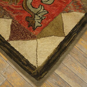Textured, looped rug corner with red, tan, and dark geometric shapes rests on distressed wood flooring.