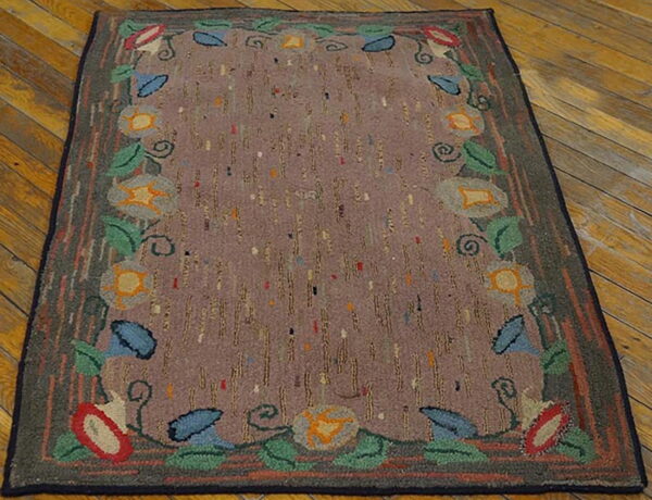 A primitive floral rug with a multicolored border framing a speckled aubergine field on distressed hardwood floors.