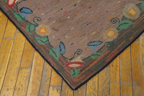 Mauve hooked rug corner with multicolored speckles, a thick floral border, and distressed yellow wood flooring.