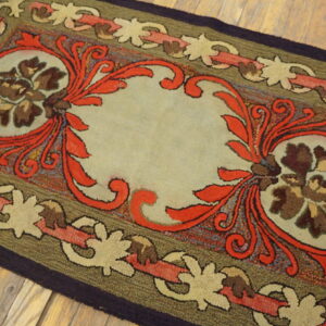 Hooked rug with a beige center, red scrollwork, and olive green, linked floral borders on wood planks.
