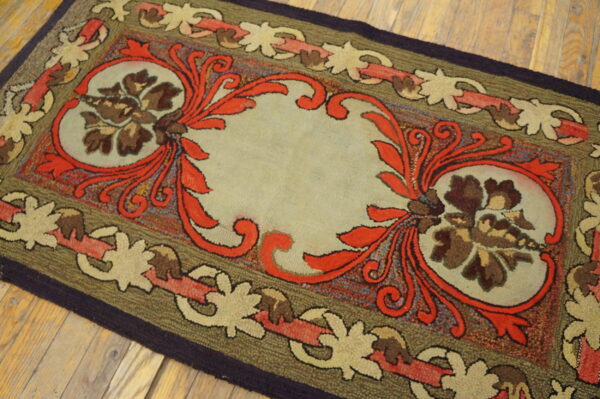 Hooked rug with a beige center, red scrollwork, and olive green, linked floral borders on wood planks.
