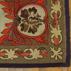 Ornate hooked rug corner featuring orange scrolls, brown florals, and an olive border on worn wood planks.
