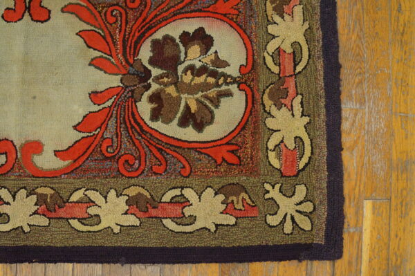 Ornate hooked rug corner featuring orange scrolls, brown florals, and an olive border on worn wood planks.