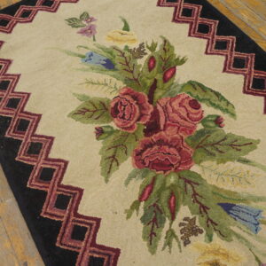 Beige hooked rug with red, green, and blue floral design and black stepped geometric border on wood floor.