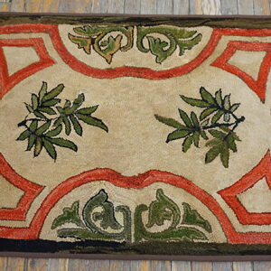 A textured rug in beige, rust, and olive green with scrolling borders and central foliage on wood flooring.