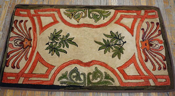 A textured rug in beige, rust, and olive green with scrolling borders and central foliage on wood flooring.