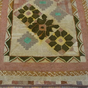 Geometric floral rug in muted green, tan, and pink with textured pile, set on distressed wooden flooring.