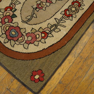 Oval hooked rug corner shows floral motifs and tan, rust, and olive borders on distressed wood floor.