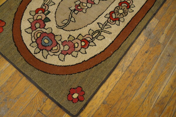Oval hooked rug corner shows floral motifs and tan, rust, and olive borders on distressed wood floor.