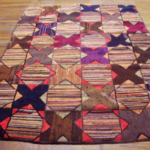 A multicolored rug featuring a geometric patchwork pattern of striped squares and dark X-shapes rests on a wooden floor.