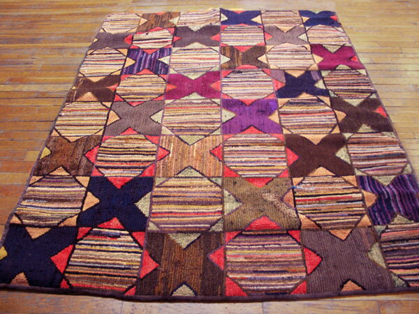 A multicolored rug featuring a geometric patchwork pattern of striped squares and dark x-shapes rests on a wooden floor.