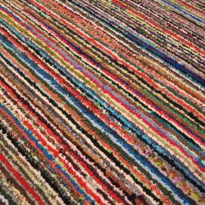 A brightly multicolored braided rag rug features narrow parallel strips of red, blue, green, and black fibers.