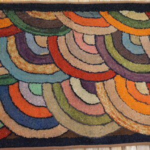 Earth-toned hooked rug with multicolored, overlapping concentric arcs and a dark border rests on wood.