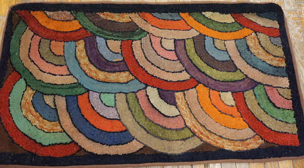Earth-toned hooked rug with multicolored, overlapping concentric arcs and a dark border rests on wood.