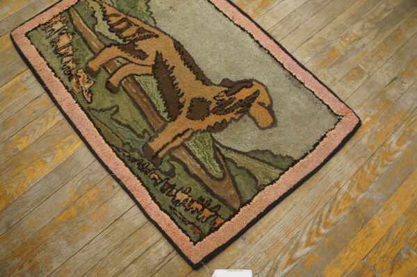 Tan and green scenic rug depicting a leaping animal, framed by a thick pink border on rustic wood flooring.