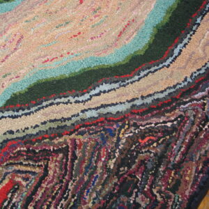 Textured rug detail with swirling bands of beige, teal, dark green, and mixed colors on a wood floor.