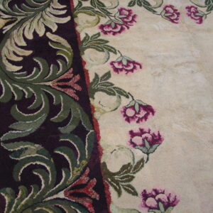 Textured rug detail showing a dark plum border with green leaves next to a cream field with pink flowers on wood flooring.