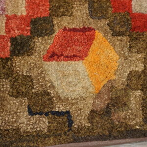 Close-up shows a hooked rug texture with tan, orange, red, and brown geometric patterns on wood flooring.