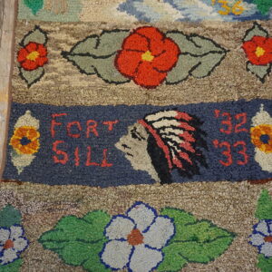 Close-up of a chunky hooked rug featuring red flowers and a dark blue band with "FORT SILL" text.