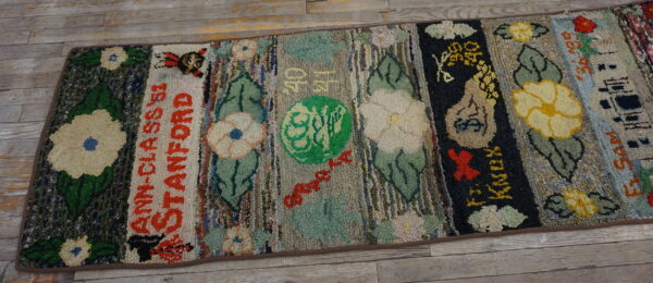 A colorful, high-pile hooked rug featuring striped panels with flowers and stitched lettering lies on wood planks.