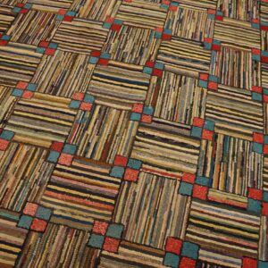 Textured diagonal grid rug with alternating horizontal and vertical multicolor stripes, punctuated by small red and teal blocks.