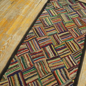Textured runner rug with multicolored striped patchwork blocks and a black border on worn wood floors.