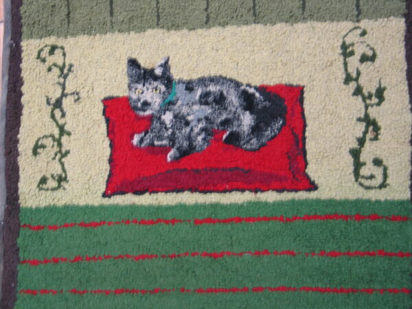 Hooked rug showing a speckled cat on a red pillow with cream, olive, and striped green borders.