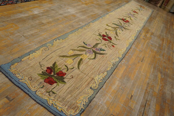 Hooked runner rug with a light background, red floral motif, and blue border, displayed on distressed wood.