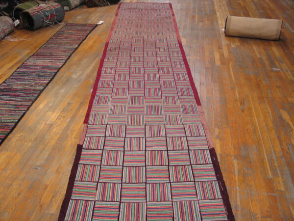 A long runner rug featuring a checkerboard pattern of vertical and horizontal multicolored stripes on wood flooring.