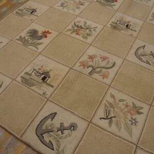 Checkerboard hooked rug in beige with squares depicting flowers, houses, and anchors rests on distressed wood flooring.