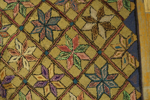 Textured olive green rug with multicolored star motifs in a diagonal grid, bordered by blue and wood floor.