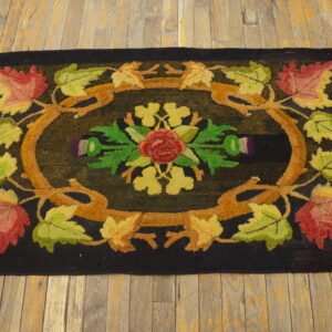 A black hooked rug featuring a central rose surrounded by colorful autumn leaves and vines on wood floors.