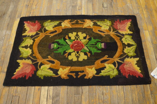 A black hooked rug featuring a central rose surrounded by colorful autumn leaves and vines on wood floors.