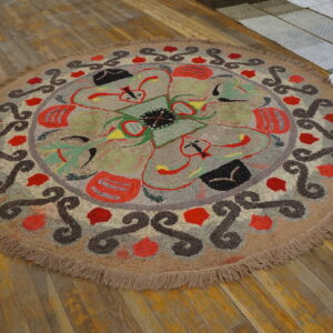 Round high-pile rug in tan, red, green, and brown abstract patterns rests on distressed wooden floorboards.