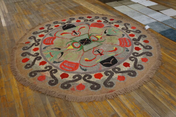 Round high-pile rug in tan, red, green, and brown abstract patterns rests on distressed wooden floorboards.