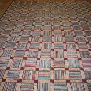 Large patchwork rug featuring a beige and maroon grid filled with multicolor striped squares on a wood floor.