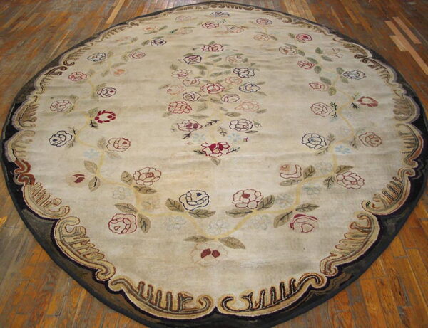An oval rug on wood flooring features a cream field with scrolling red, pink, and green floral motifs and a complex dark border.