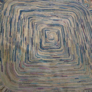 Textured rug in blues, grays, and beige featuring a central concentric square motif and scalloped edge.