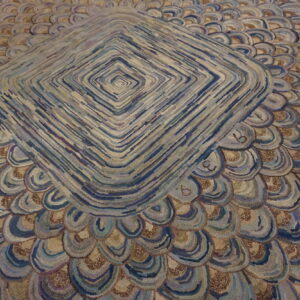 Textured rug showing a central diamond pattern surrounded by rows of blue and tan scallops.