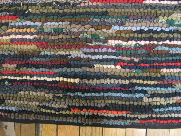 Close-up of a multi-colored hooked rag rug with dense texture and variegated horizontal rows resting on wood planks.