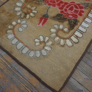 Tan loop rug with large red floral patterns and white scalloped borders rests on worn plank wood flooring.