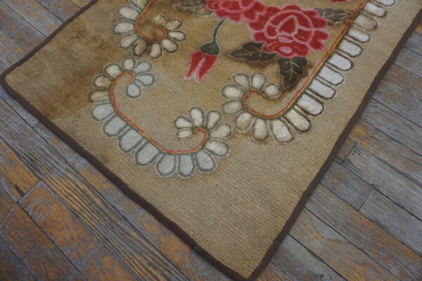 Tan loop rug with large red floral patterns and white scalloped borders rests on worn plank wood flooring.